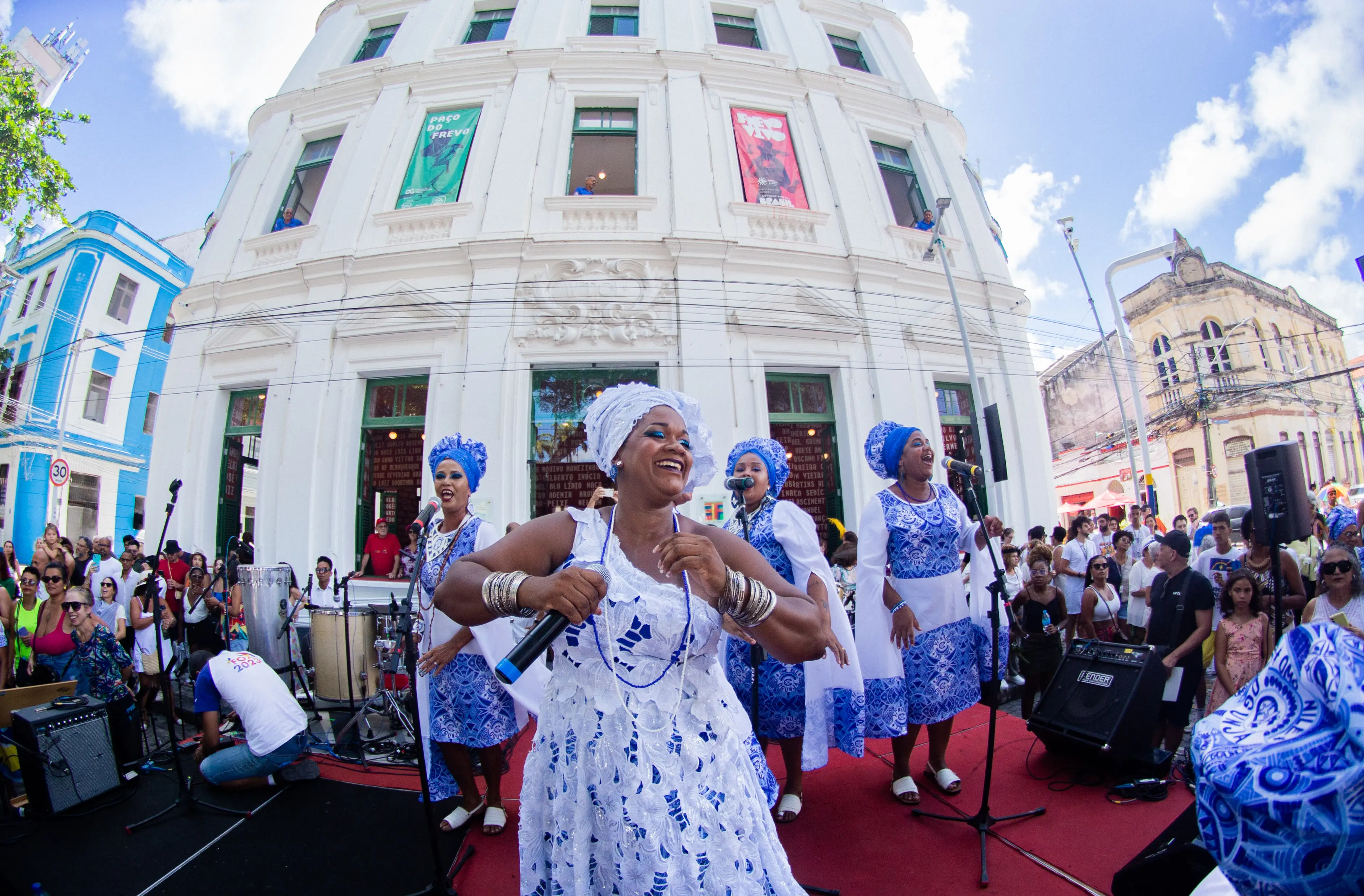 Group of women in traditional white and blue attire performing singing and dancing on a busy street, in front of a historic building, during a cultural celebration.