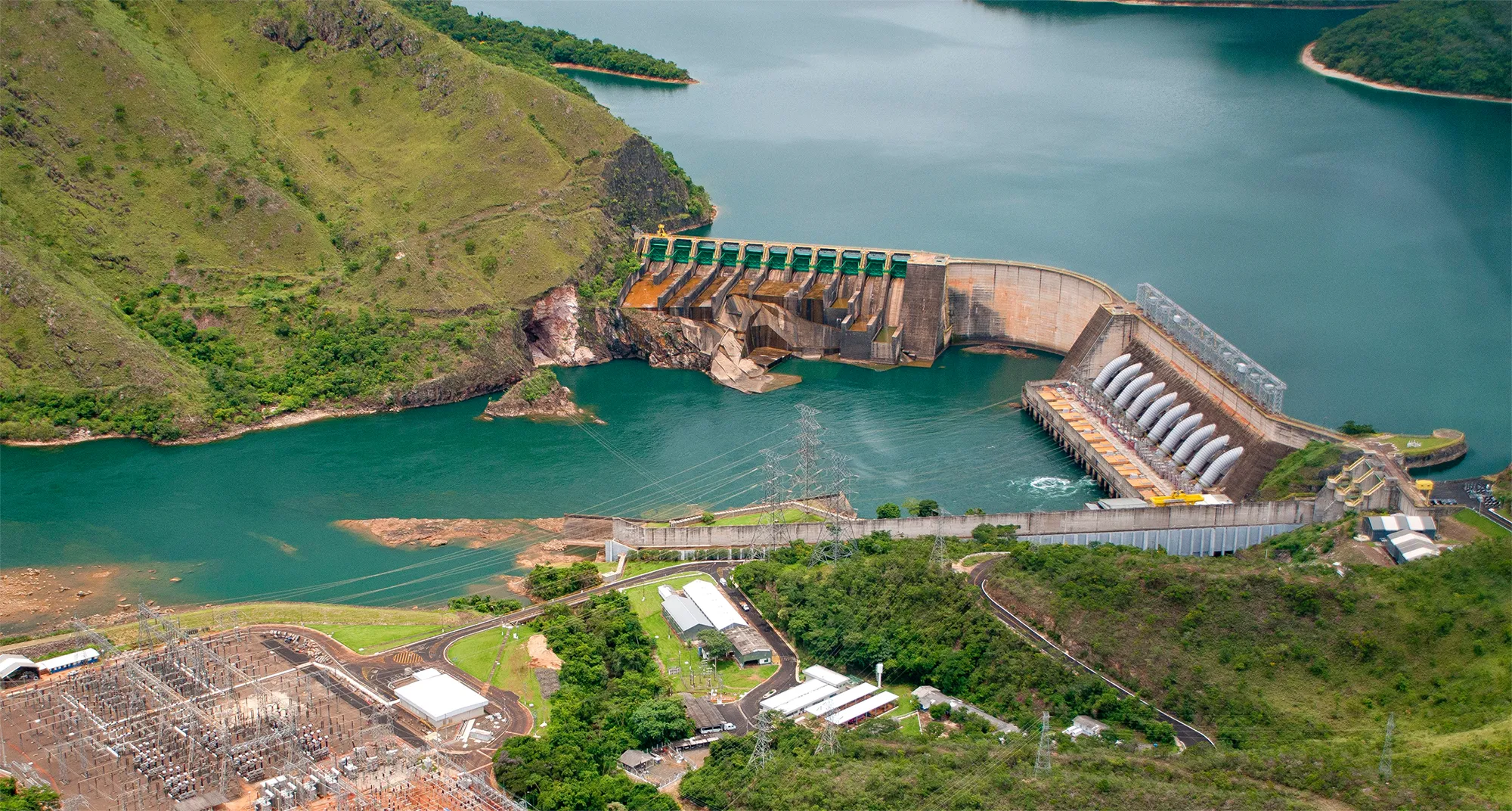 Vista aérea de uma usina hidrelétrica com barragem e turbinas geradoras de energia, rodeada por montanhas e vegetação, representando a geração sustentável de energia elétrica.