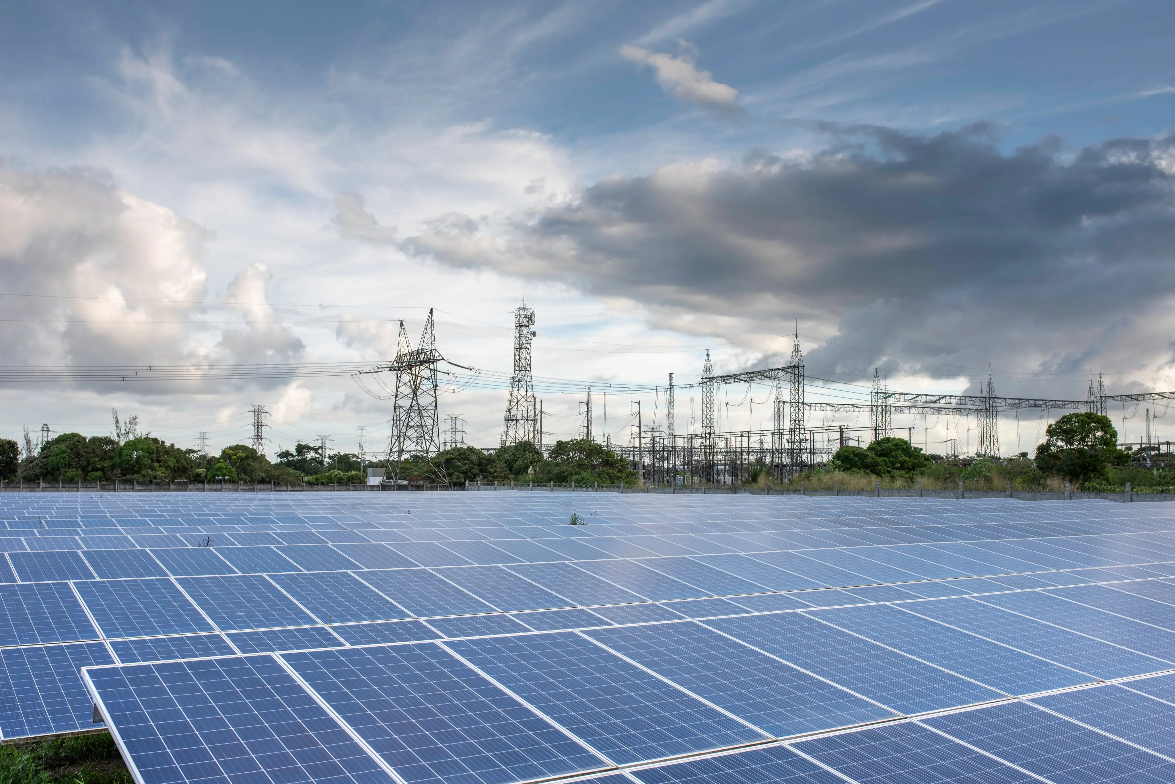 Amplo campo de painéis solares de uma usina fotovoltaica em primeiro plano, com uma subestação e torres de transmissão de energia ao fundo, sob um céu com nuvens dramáticas.