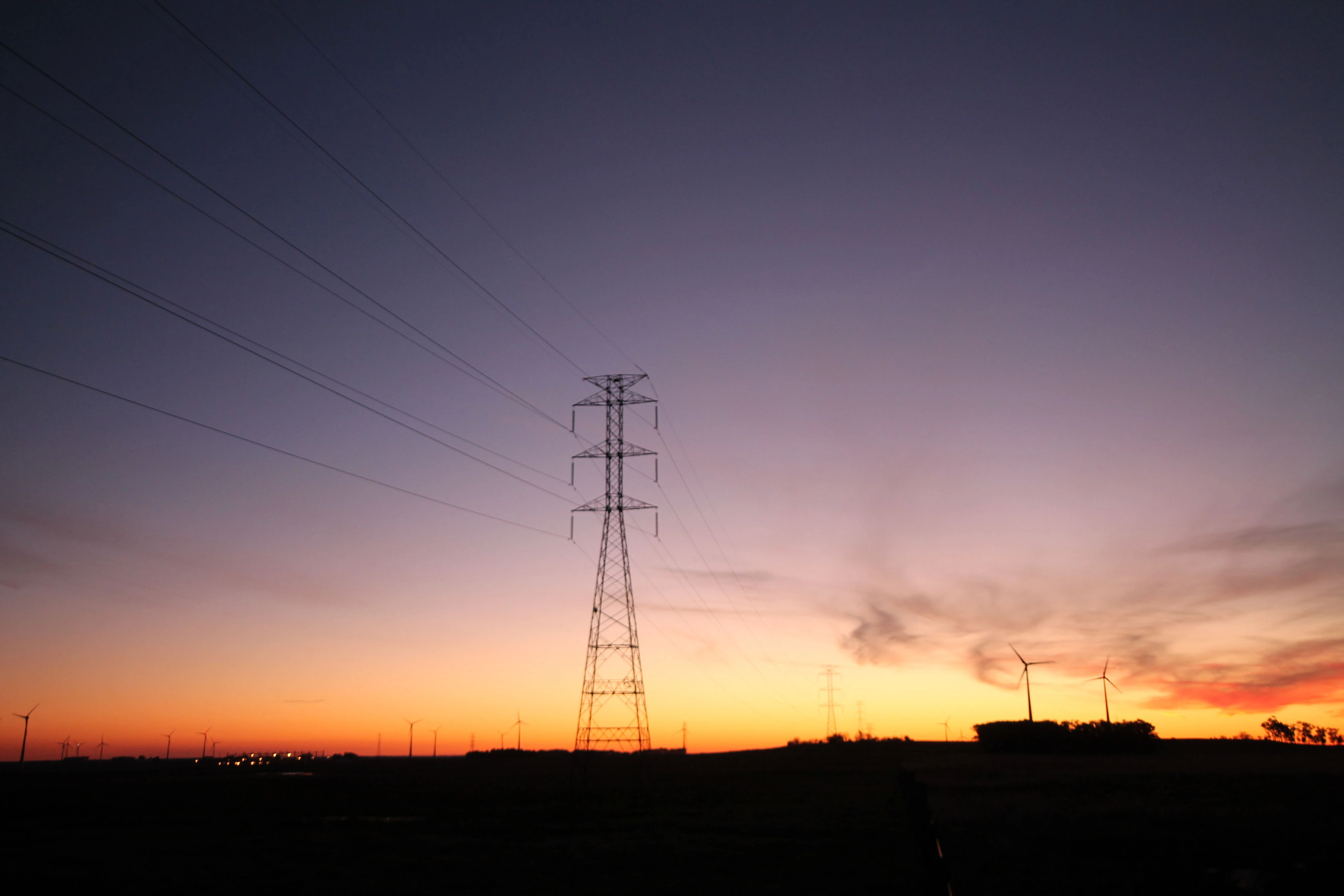 Paisagem ao entardecer com a silhueta de uma torre de transmissão de energia no centro e várias turbinas eólicas ao fundo. O céu exibe um gradiente de cores do laranja ao roxo.